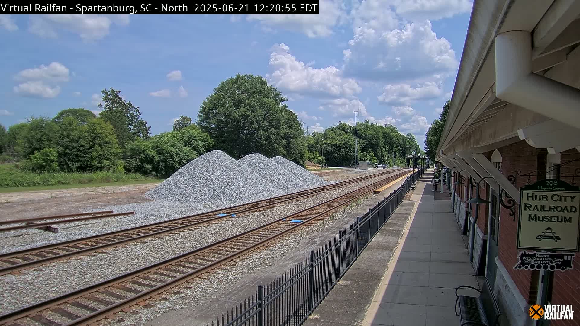 Three piles of gravel sit beside railroad tracks next to a station platform under a partly cloudy sky.
