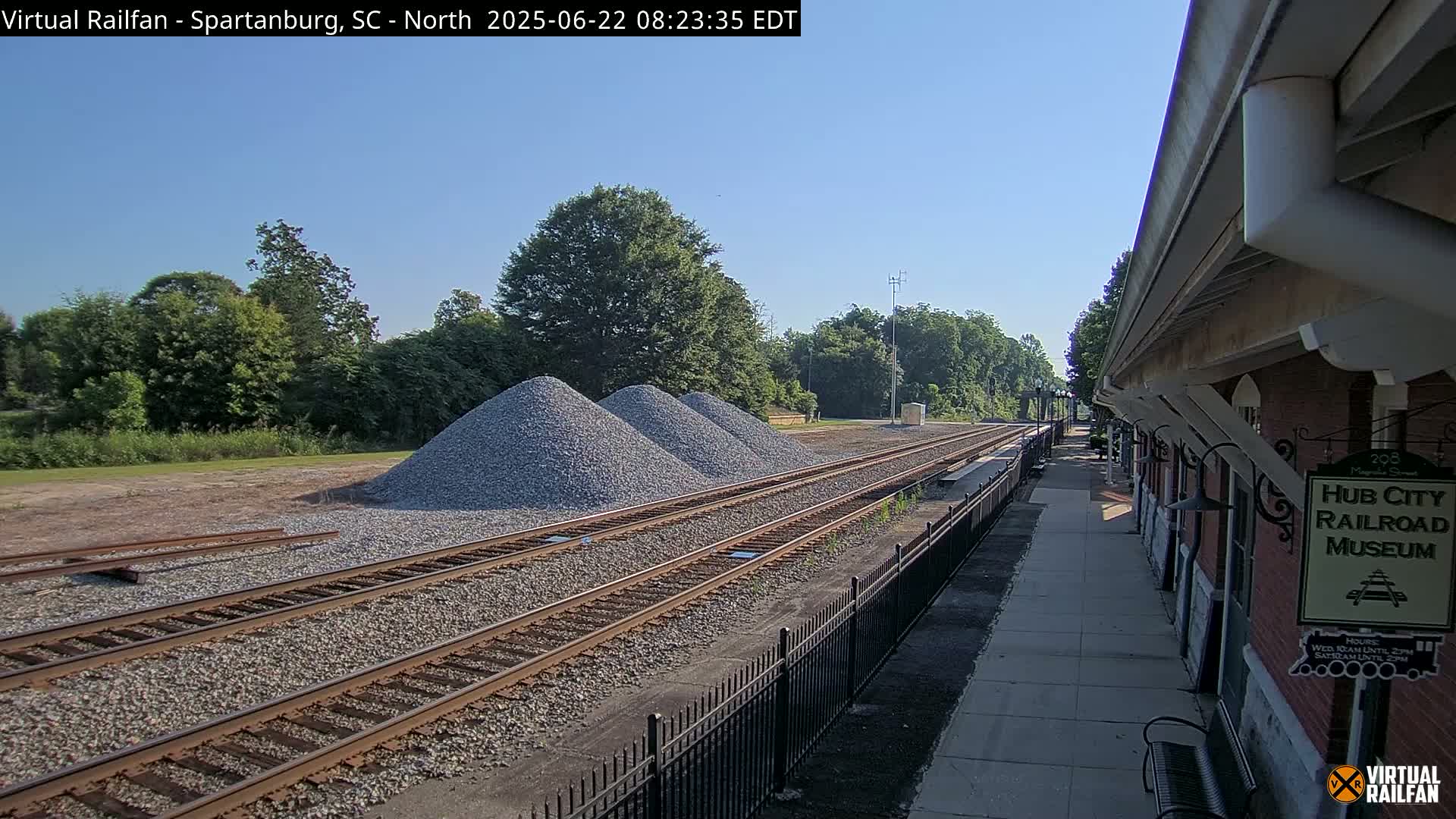 Under a clear sky, railroad tracks extend from a brick building alongside piles of gravel toward a line of trees.