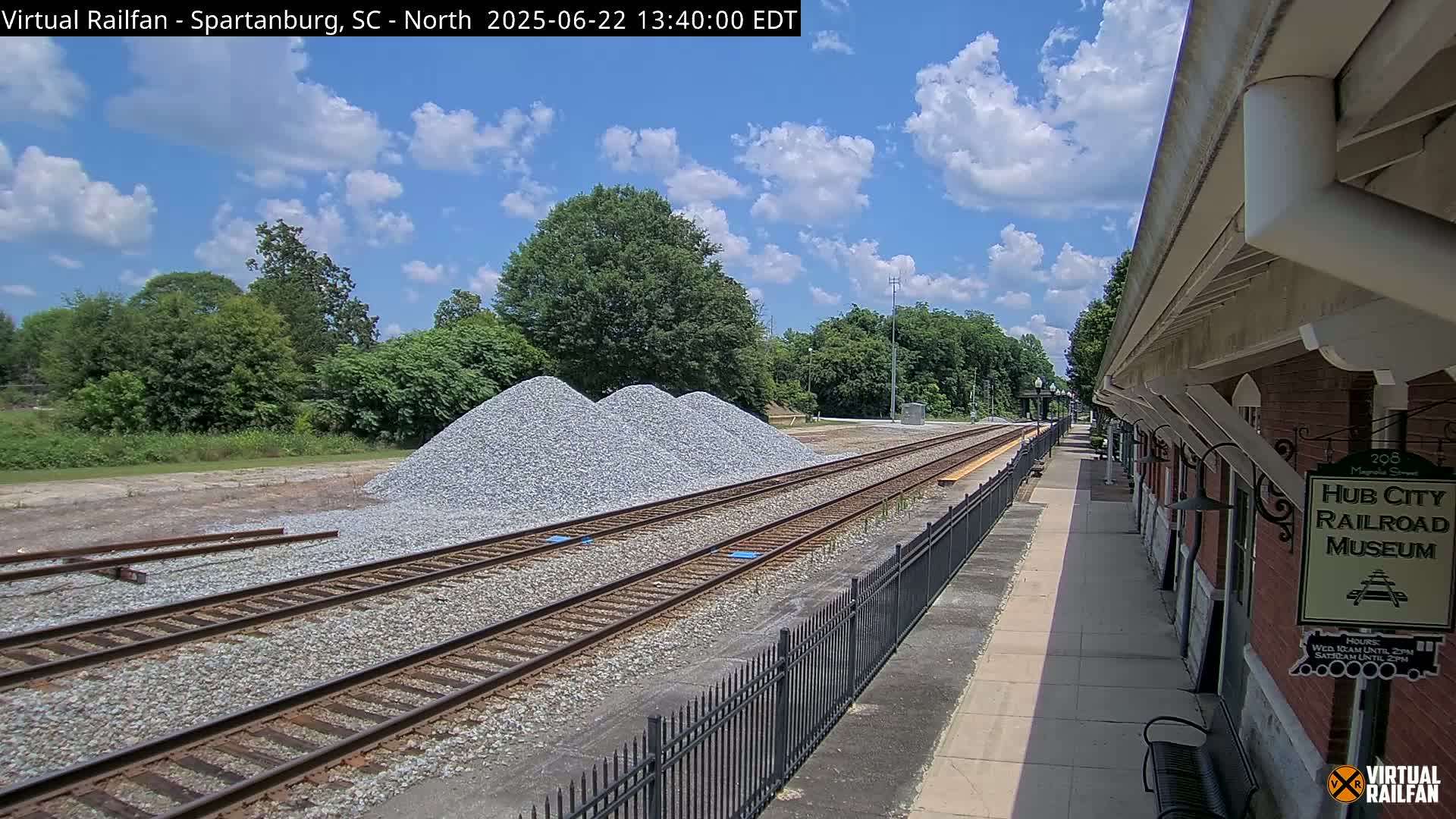 A partly cloudy day reveals a railroad track with piles of gravel beside it, next to a brick railroad museum building.