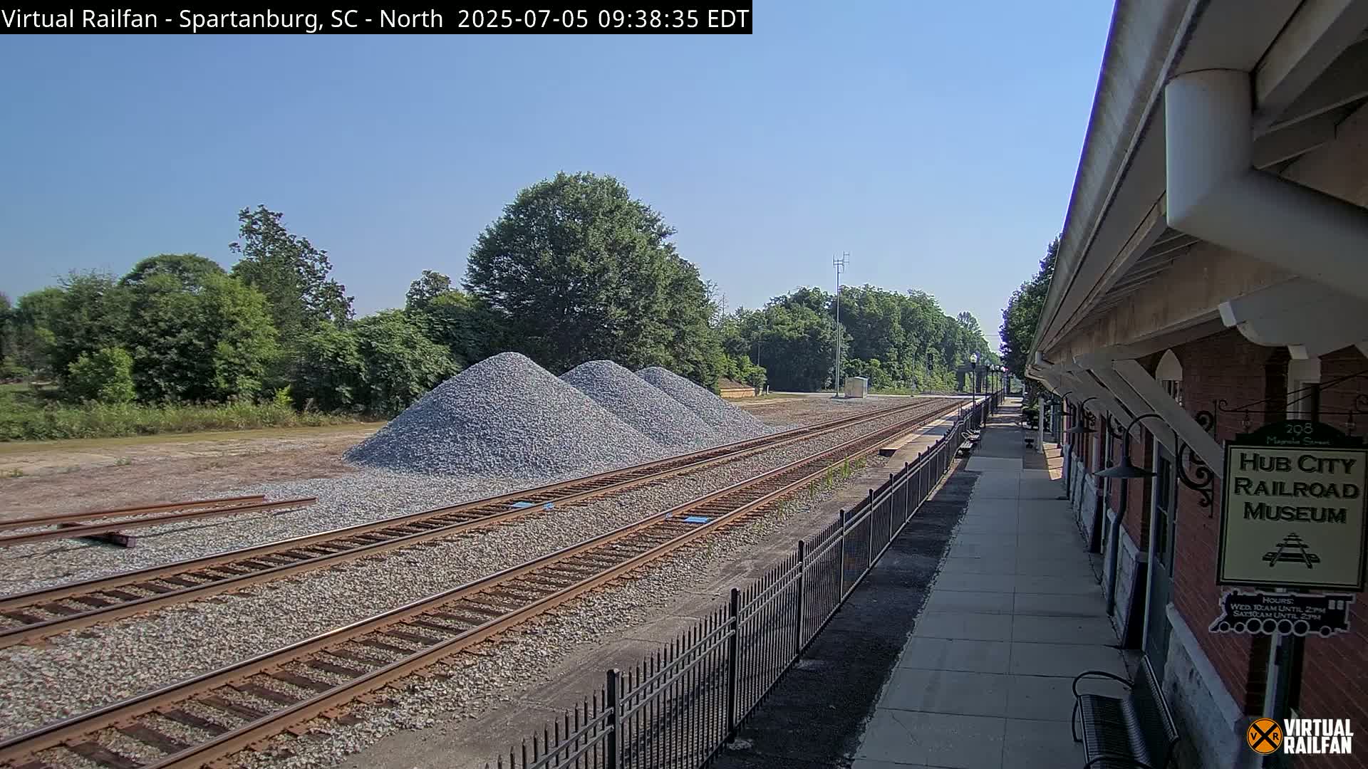 Several piles of gravel sit beside railroad tracks next to a brick building on a sunny day.