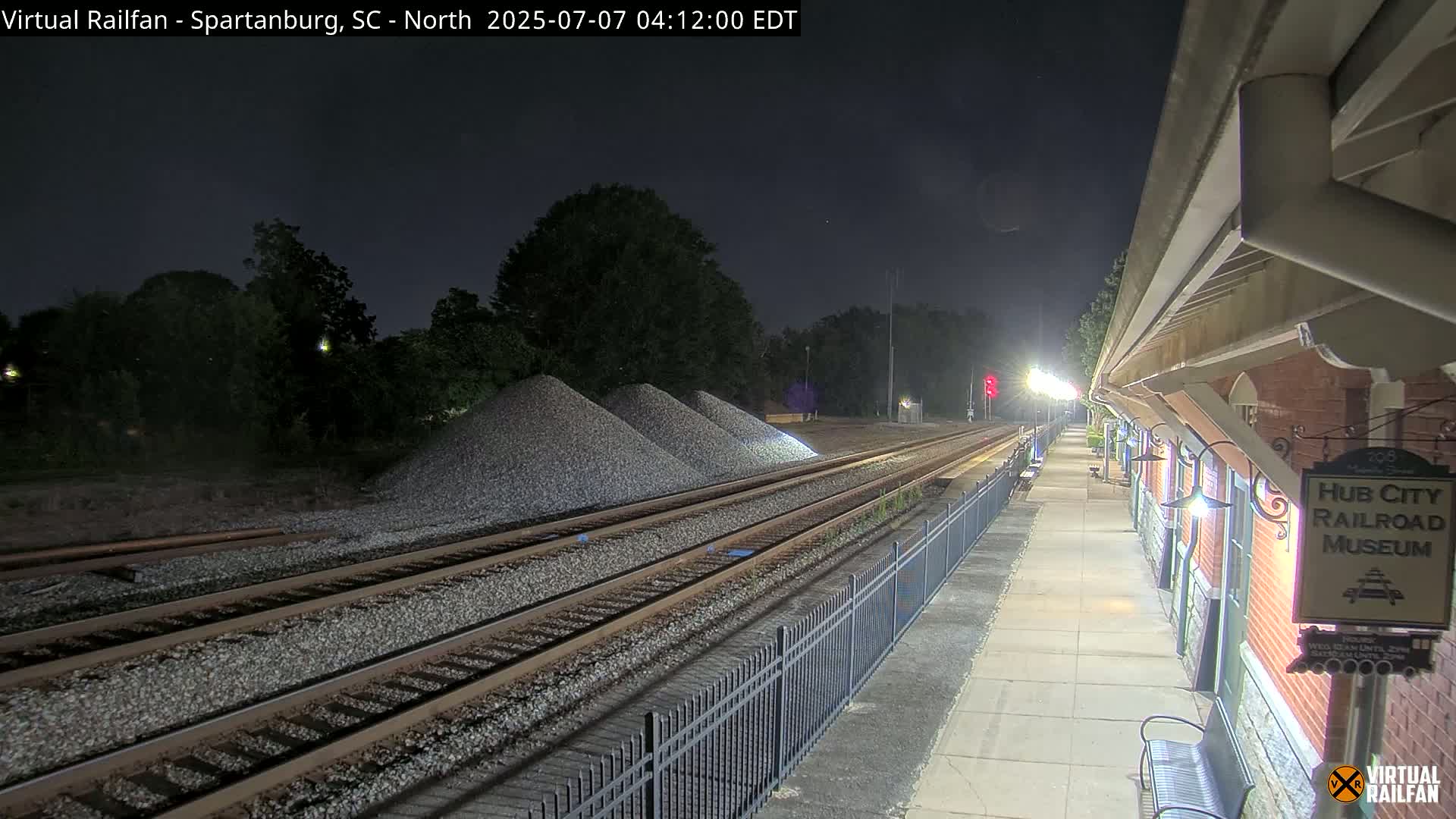 At night, under a dark sky, several sets of train tracks extend from a lit train platform toward piles of gravel next to a line of trees.