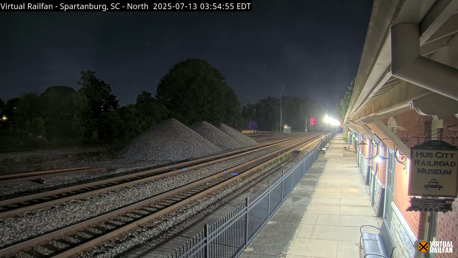 Under a dark night sky, railroad tracks extend from a nighttime train station toward piles of gravel.