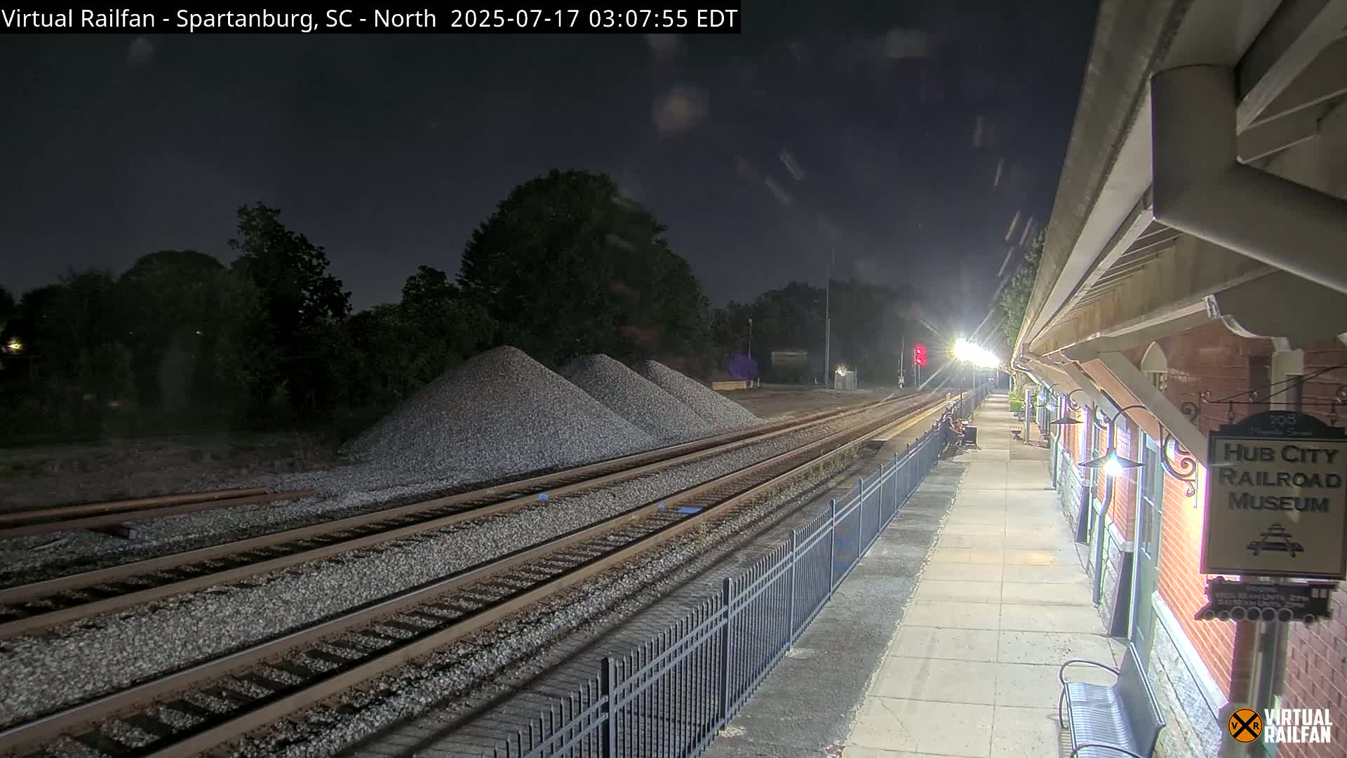 At night, a train station platform with a brick building and several piles of gravel next to train tracks is illuminated under artificial light.