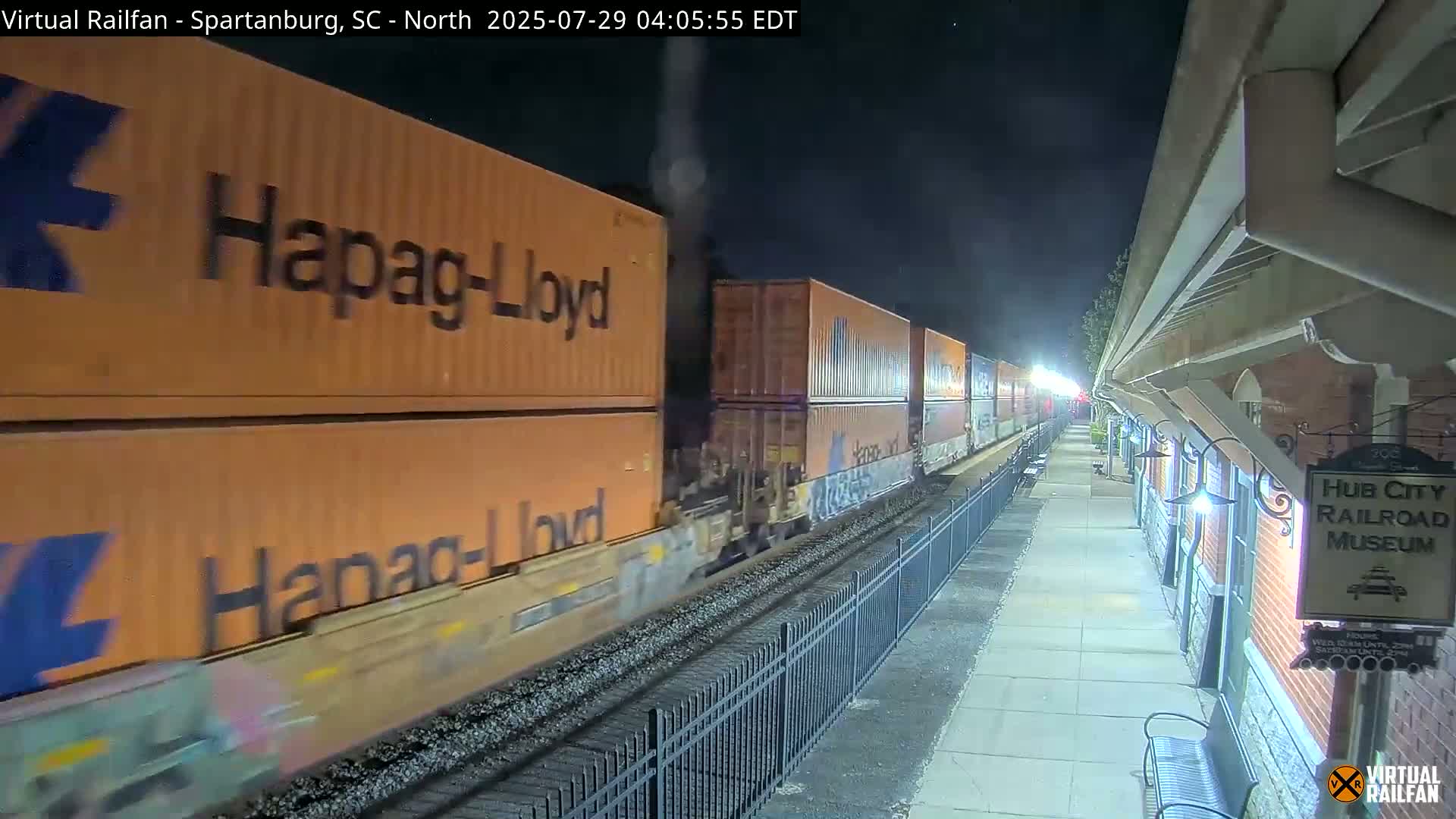At night, a long freight train carrying orange shipping containers passes by a railroad museum platform.