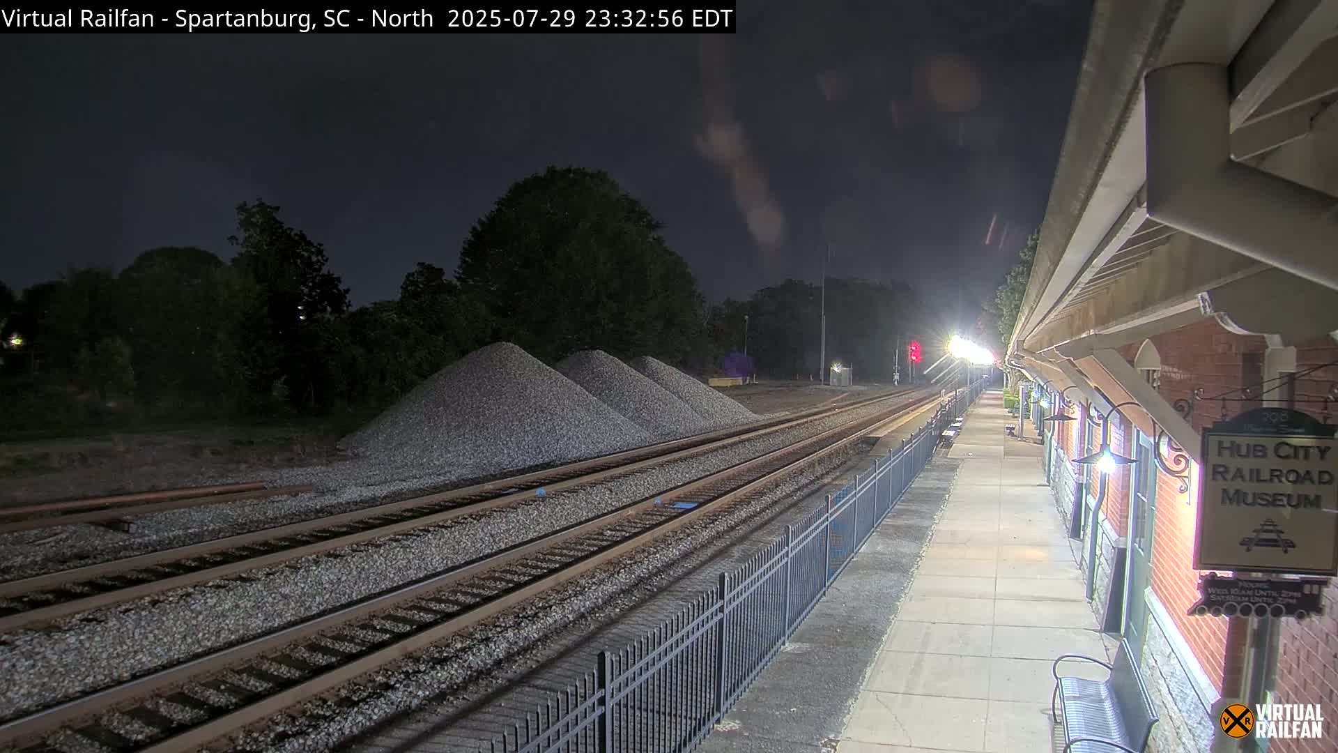 At night, a long freight train carrying orange shipping containers passes by a railroad museum platform.