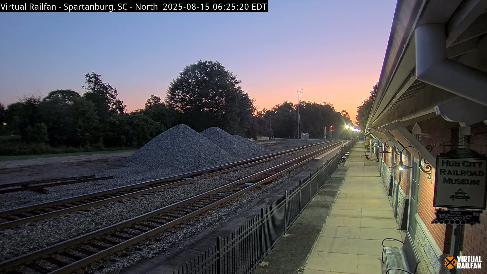 At night, a long freight train carrying orange shipping containers passes by a railroad museum platform.