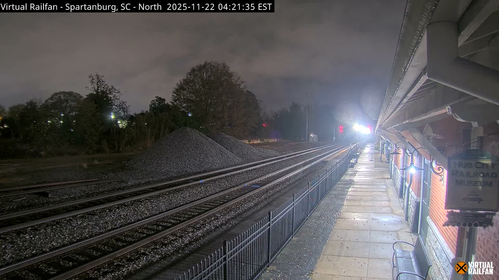 On an overcast and damp night, multiple train tracks run alongside an illuminated station platform and a brick building with an awning, with piles of gravel next to the tracks and bright lights visible in the distance.