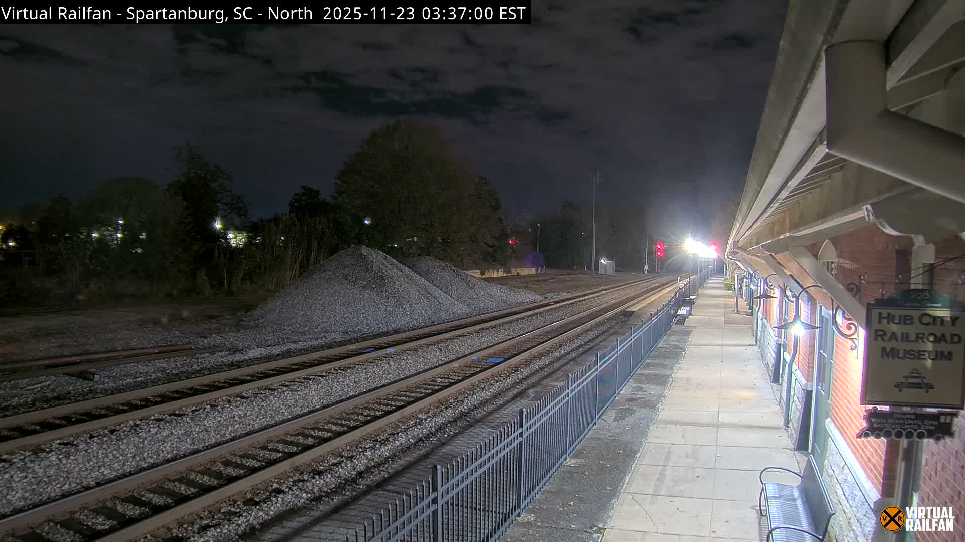 A brightly lit railway station platform with benches and an overhead shelter stands beside multiple tracks and large piles of gravel, all under a cloudy night sky.