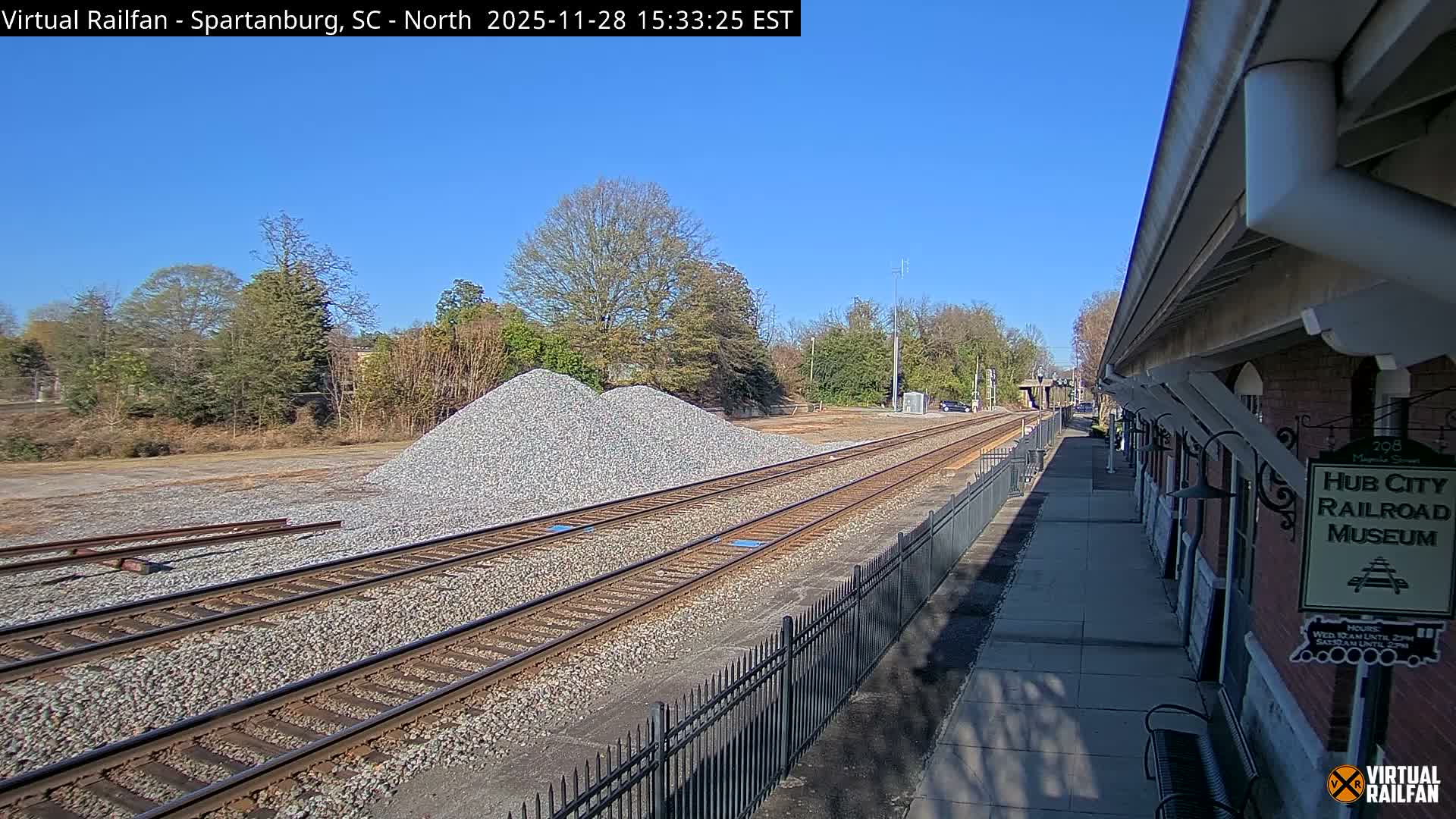 Under a clear and sunny sky, multiple railroad tracks run alongside a large pile of gravel and a long brick building with a covered platform.