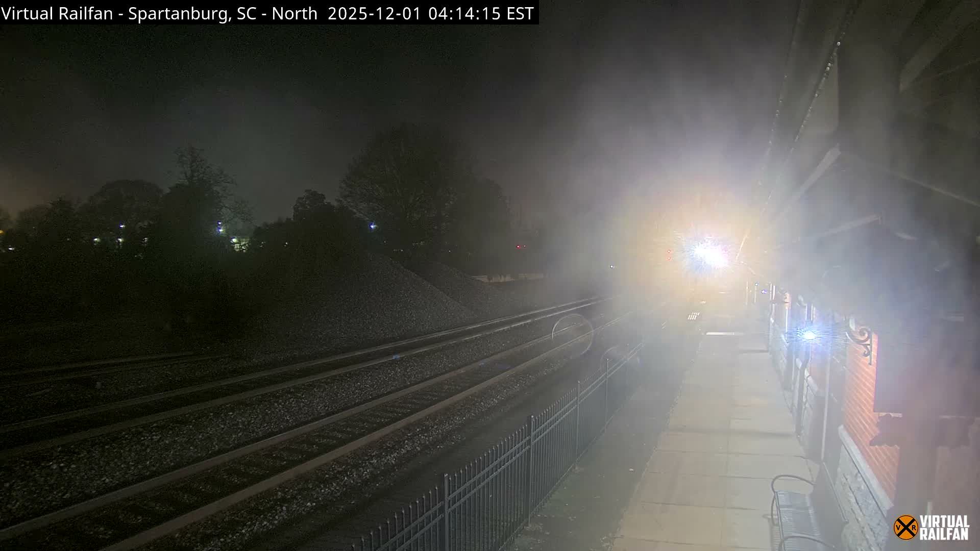 A train with bright headlights approaches a station platform and multiple tracks on a foggy night, casting light on the surrounding area.