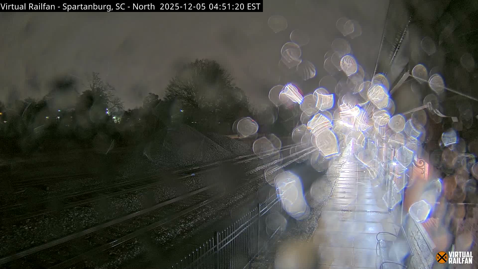A rainy nighttime scene reveals multiple train tracks stretching into the distance alongside a dimly lit platform with benches, all viewed through a heavily rain-streaked lens.