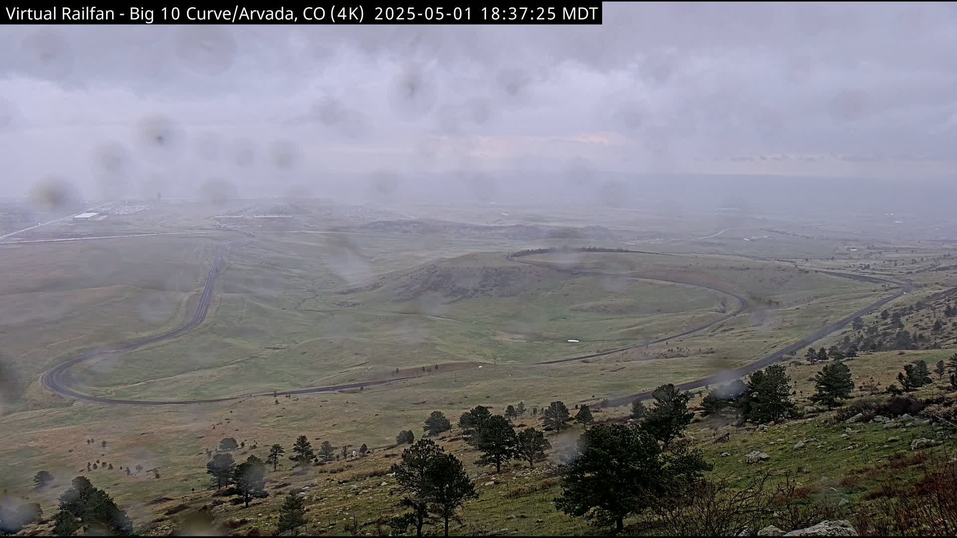 A winding road curves through a hilly, grassy landscape under a cloudy, rainy sky.
