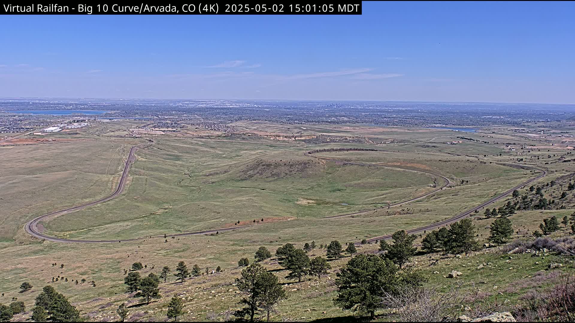 A high-angle view on a clear day shows a winding road curving through a grassy landscape, with a distant city skyline visible on the horizon.