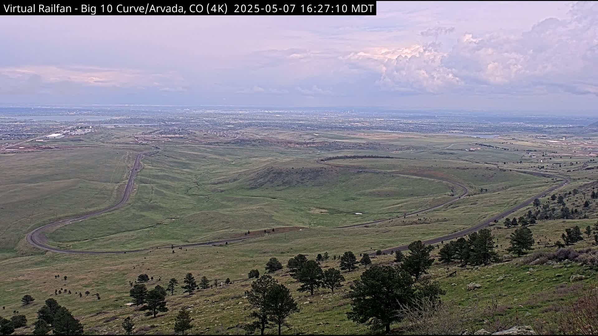 A winding road curves through a sprawling green landscape under a partly cloudy sky, with a distant city visible on the horizon.