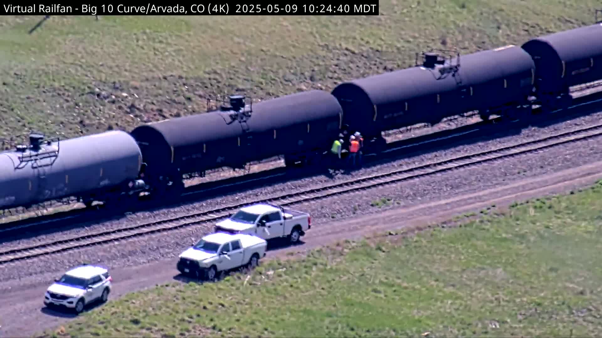 A long freight train of tanker cars is stopped on railroad tracks next to a grassy area, with several white pickup trucks and a few people standing nearby.