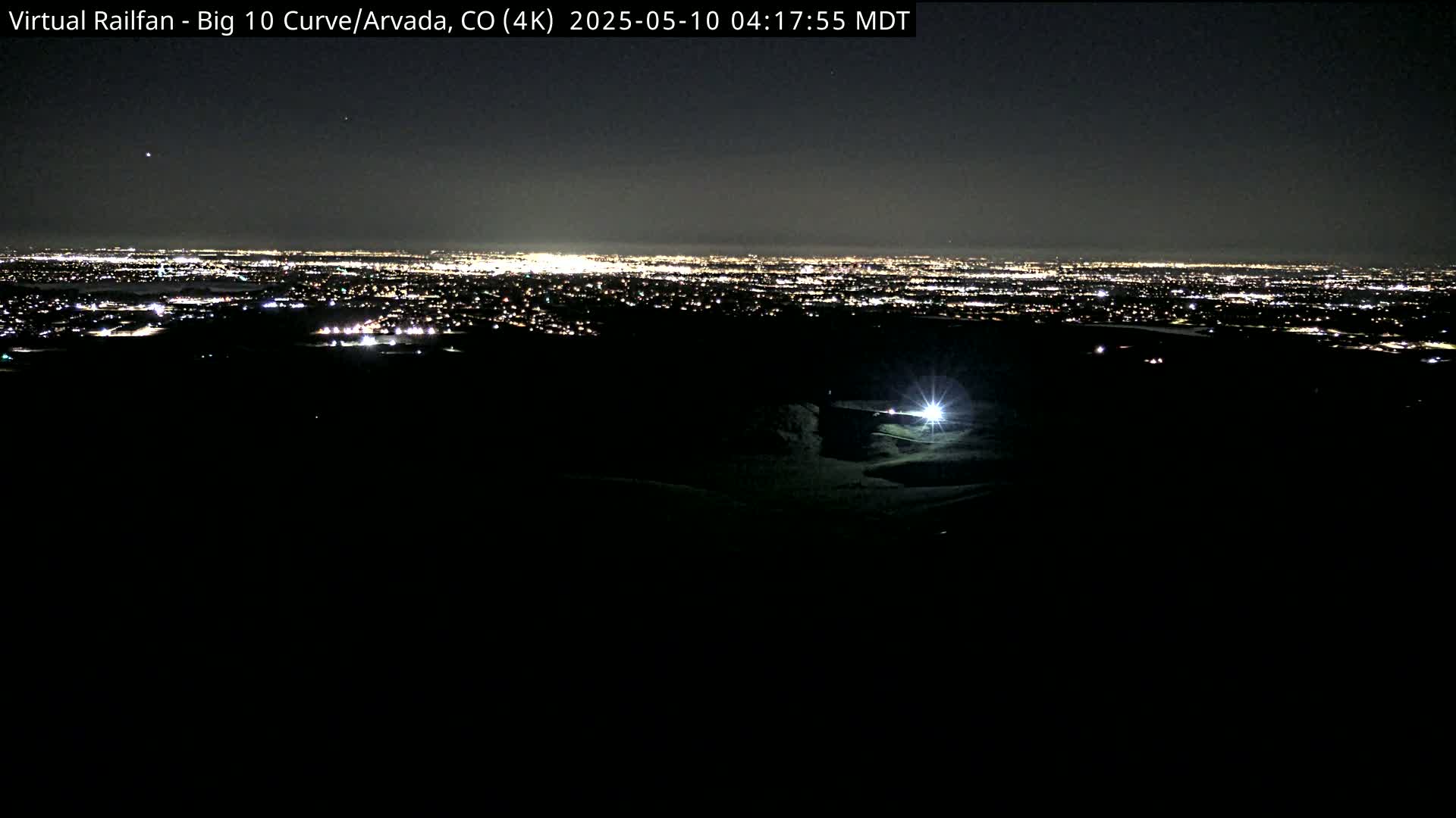 A nighttime view shows a brightly lit city in the distance, with a single light source visible in the dark foreground under a clear sky.