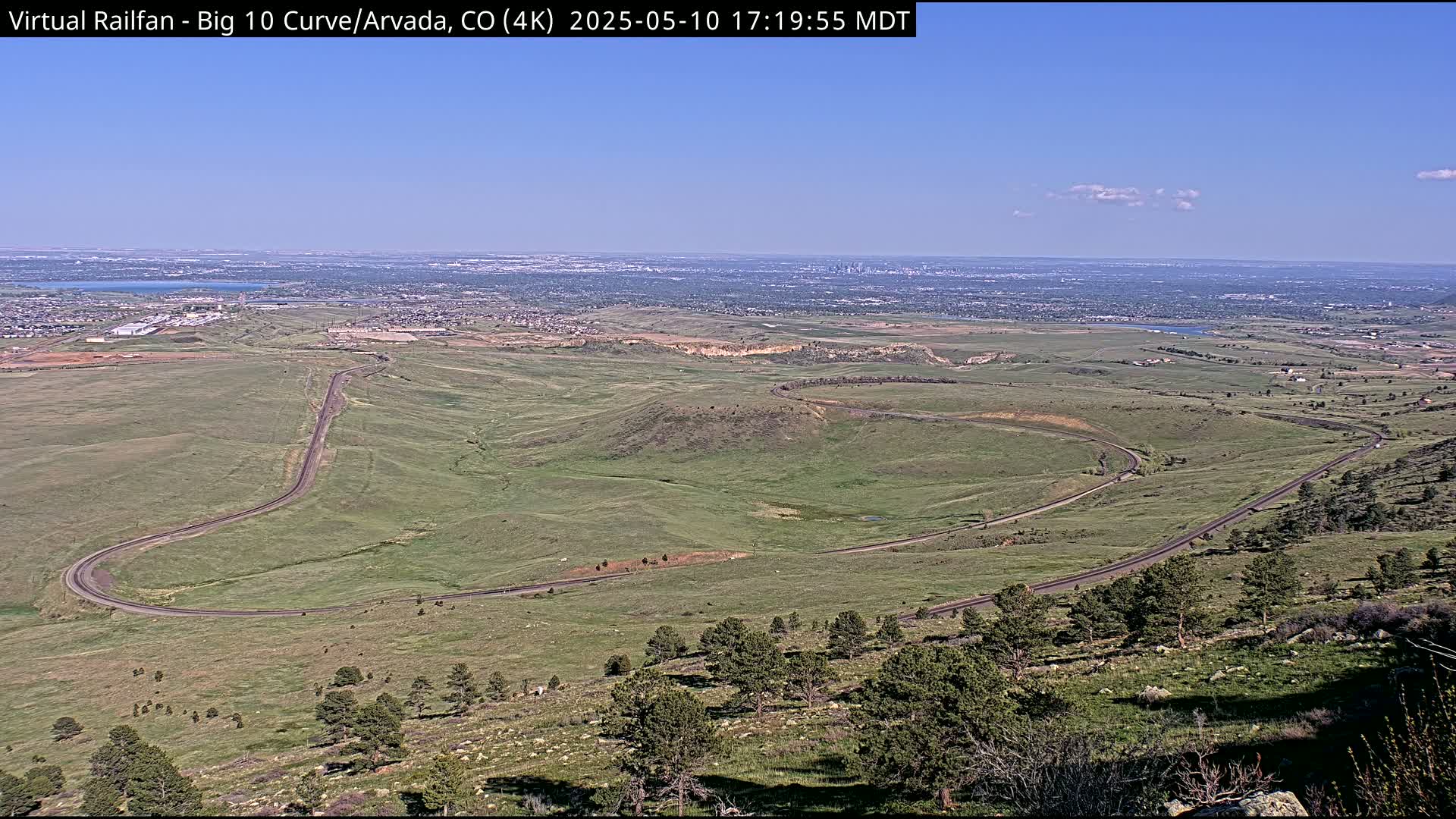 A winding road curves through a grassy landscape, overlooking a distant city under a clear blue sky.