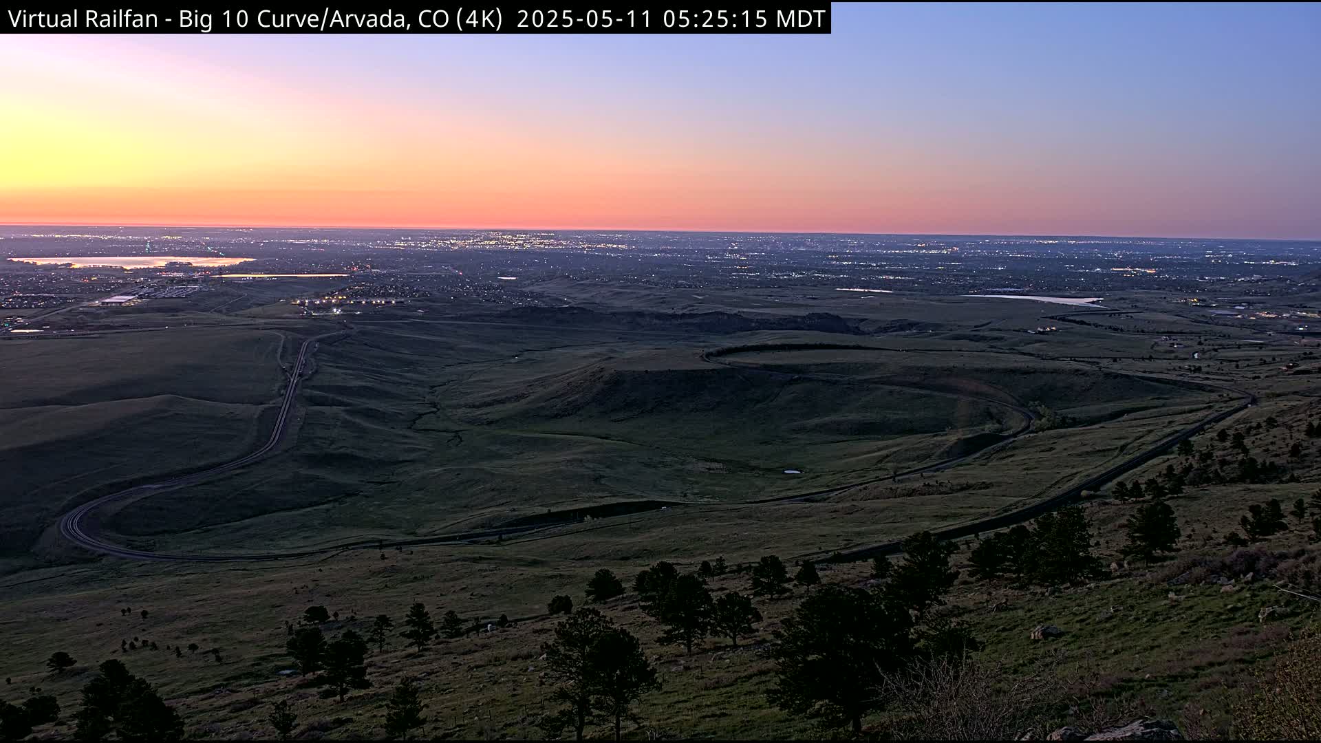 A winding road curves through a hilly, dark green landscape at sunrise, overlooking a distant city illuminated by early morning lights.