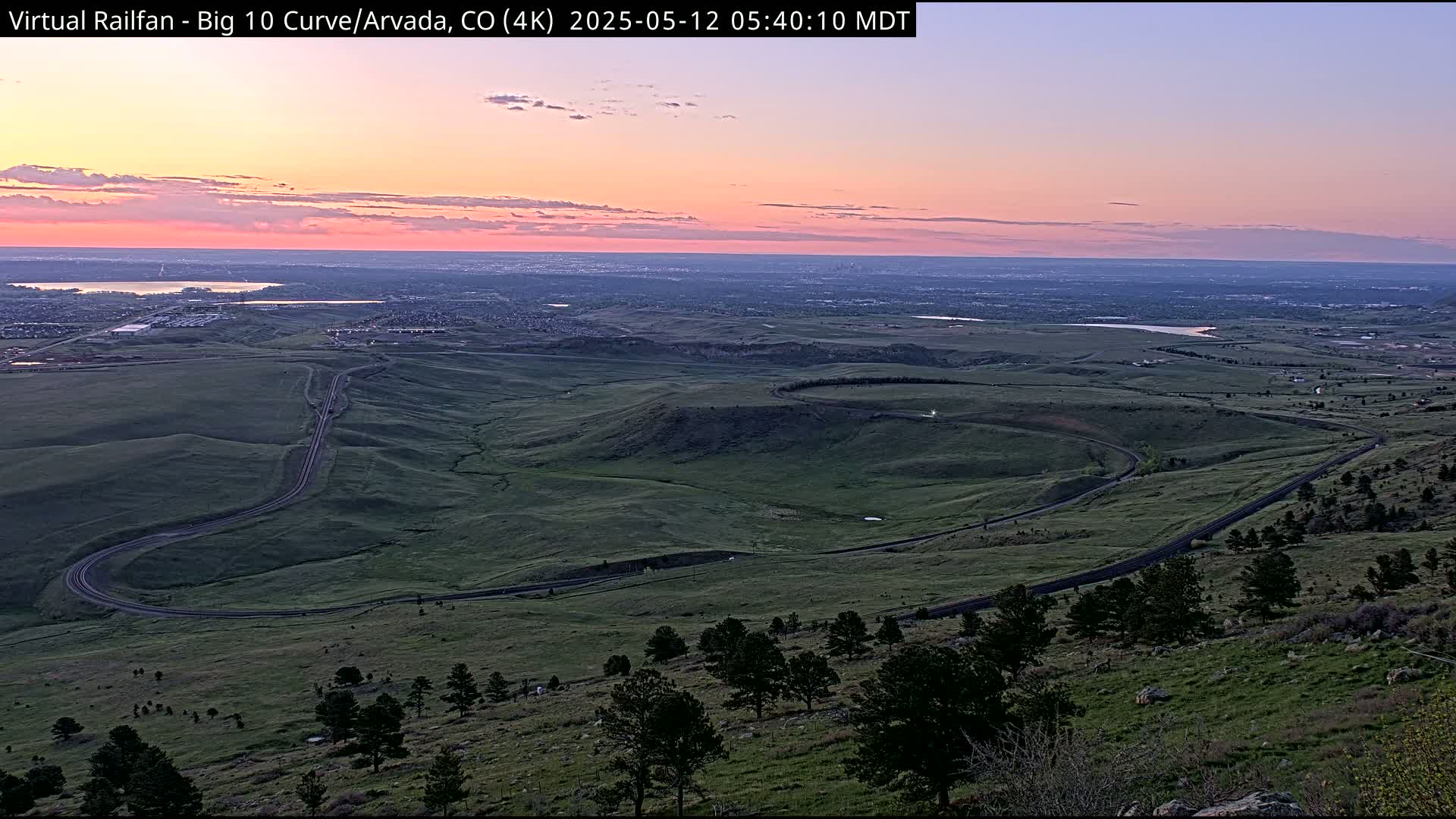 A winding road curves through a green, hilly landscape under a pastel sunset sky over a distant city.