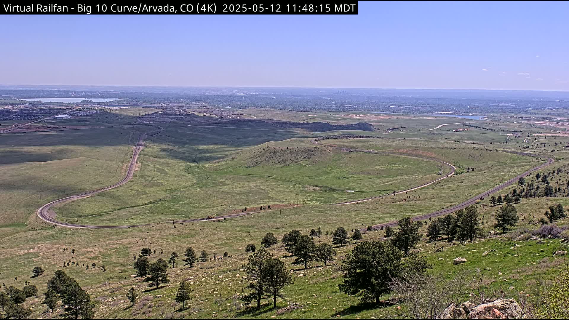 A winding road curves through a grassy hillside under a clear blue sky, with a distant city visible on the horizon.