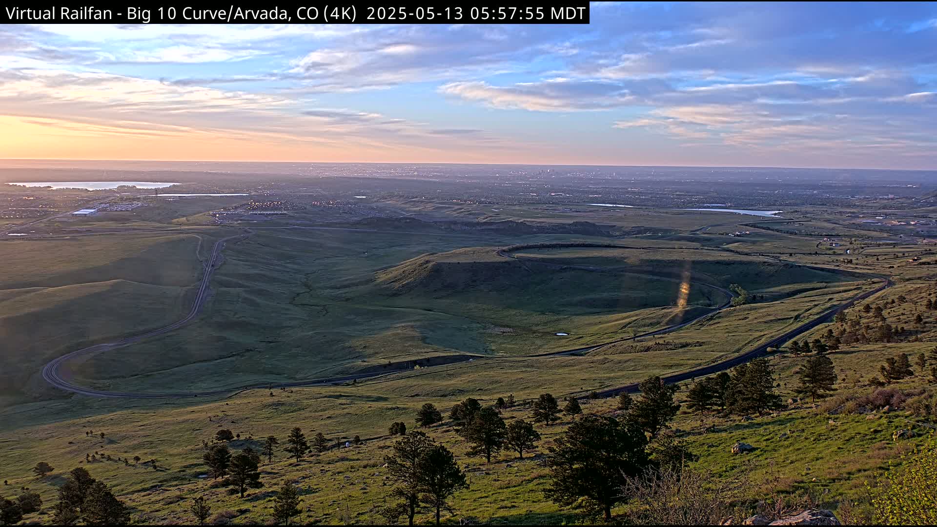 A winding road curves through a grassy landscape at sunrise, with a distant city skyline visible under a mostly clear sky.