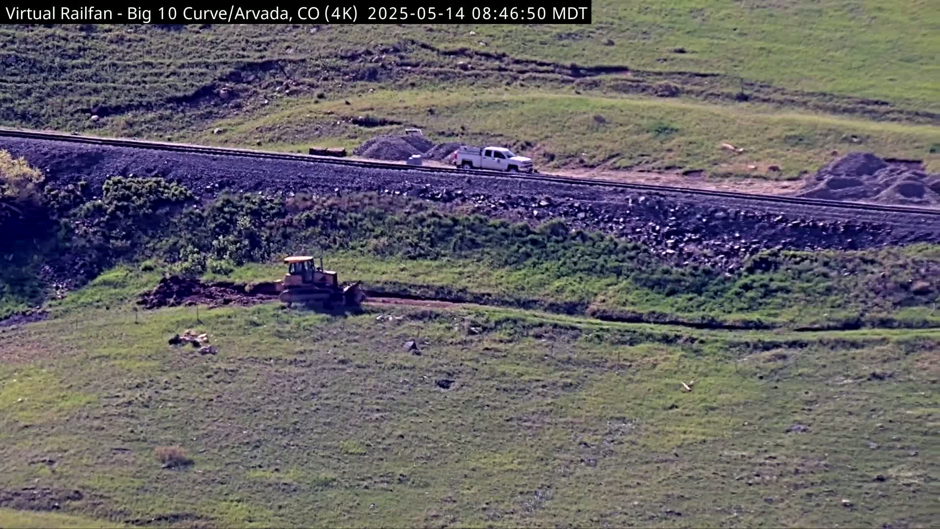A bulldozer and a pickup truck are working near a railway line on a sunny day in a grassy field.