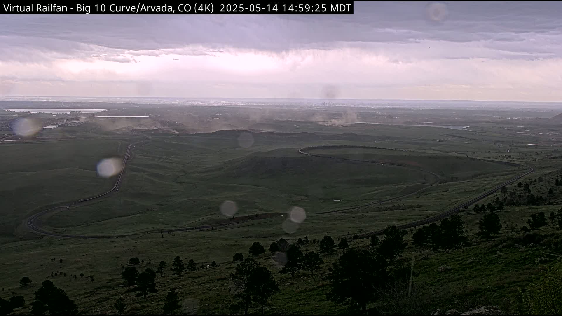 A winding road curves through a green, hilly landscape under an overcast sky, with rain visible on the camera lens and a distant city skyline faintly visible in the background.