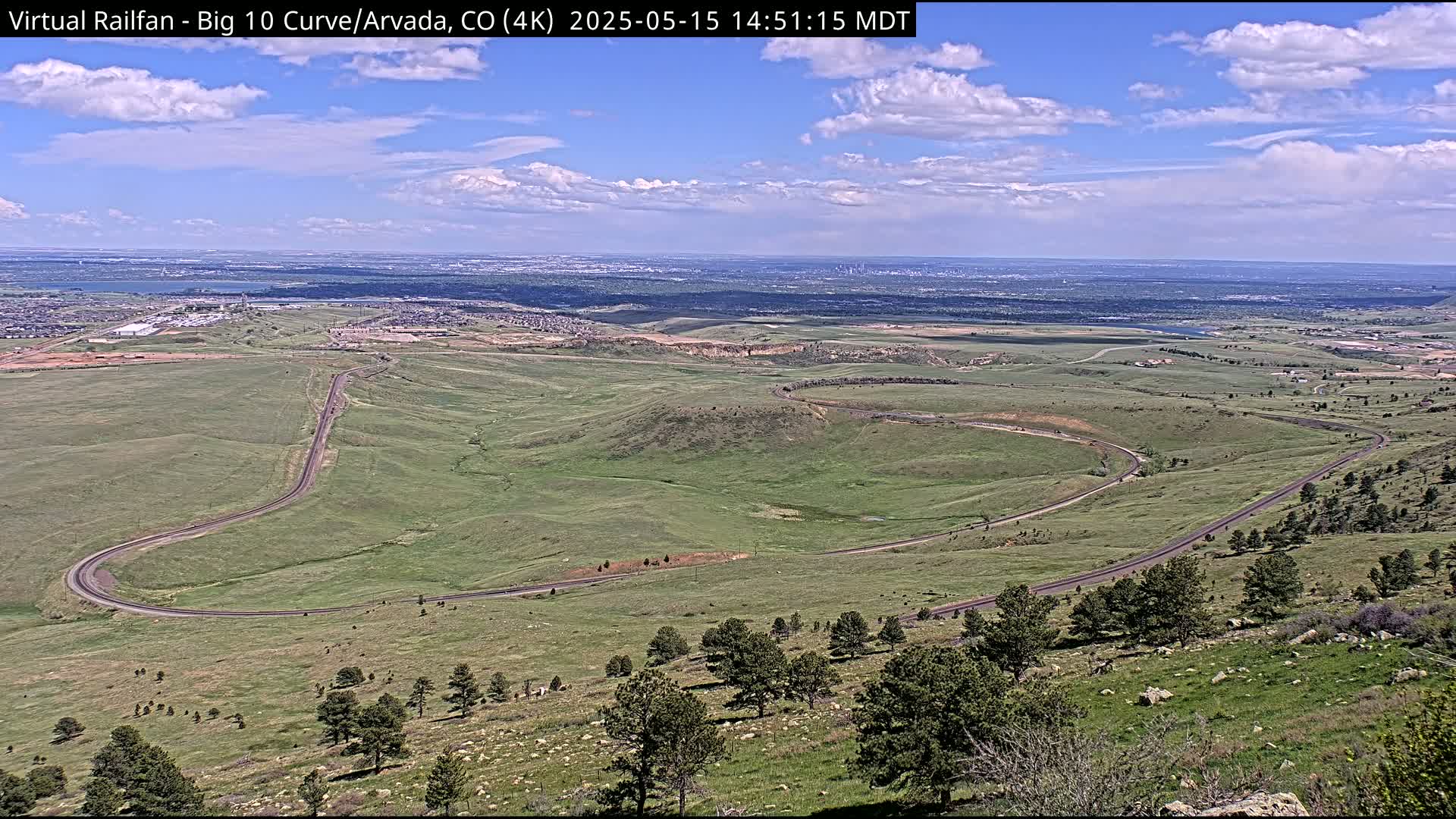 A winding road curves through a grassy landscape under a partly cloudy sky, with a distant city visible on the horizon.