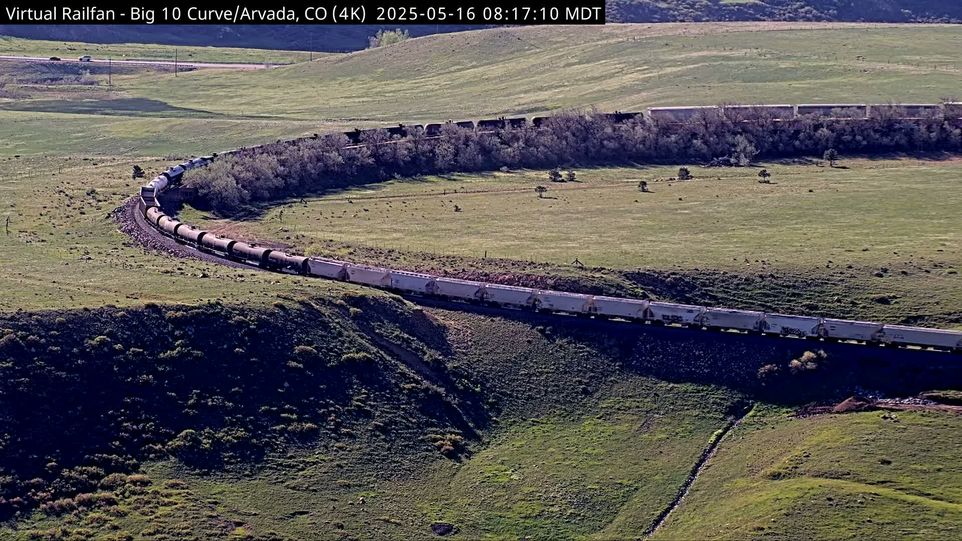 A long freight train curves around a hillside on a sunny day in a grassy, open landscape.