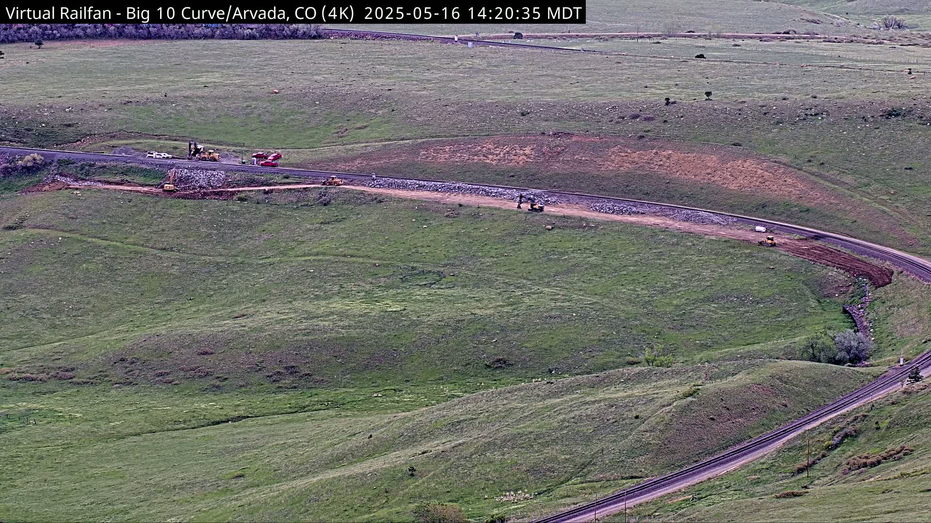 Construction equipment is working on a road and railway curving through a grassy field under a mostly clear sky.