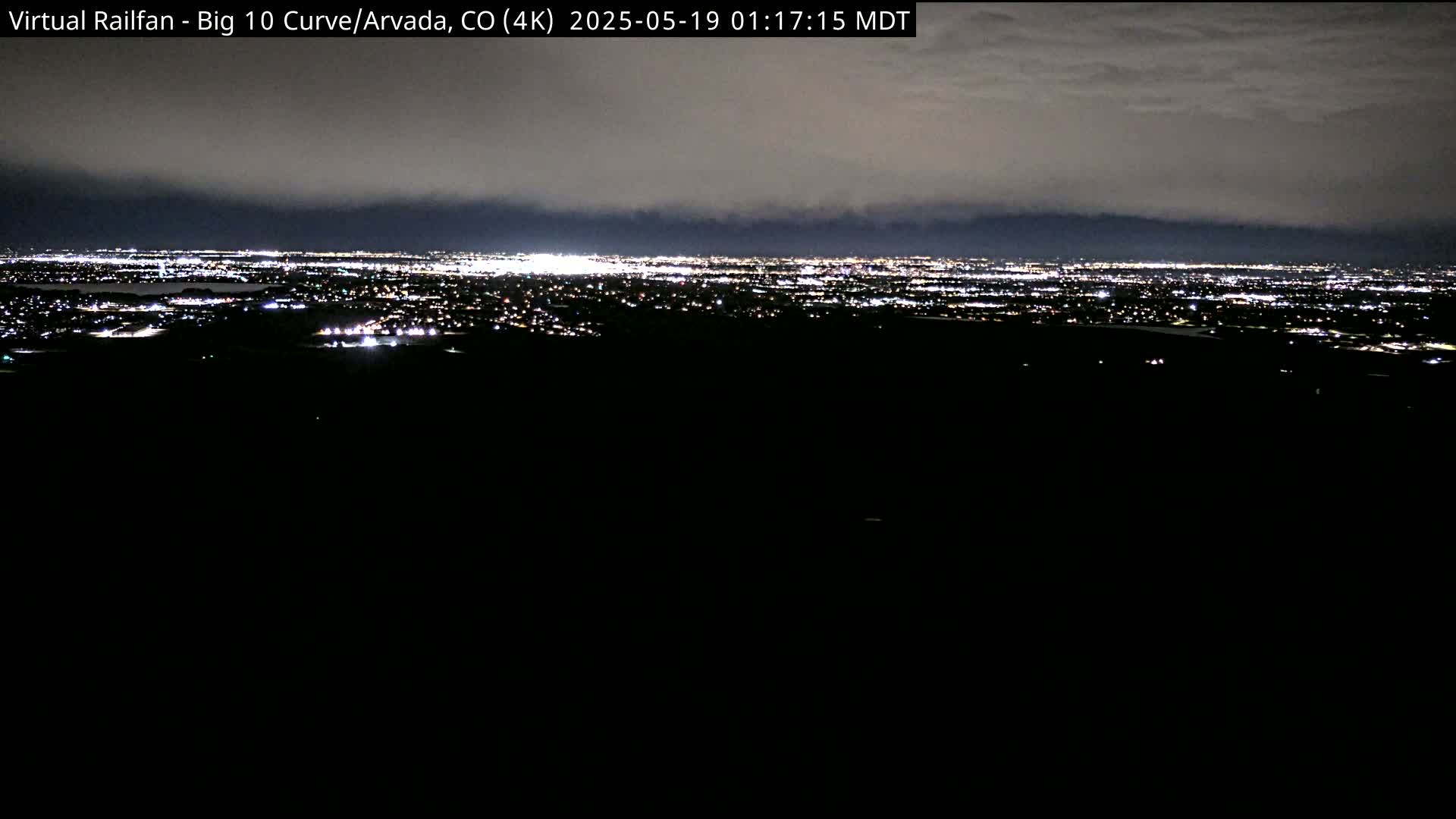 A nighttime, overcast view shows a sprawling city illuminated below, stretching to a dark mountain range in the distance.