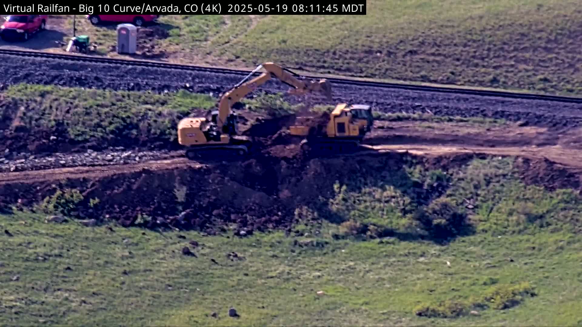Two yellow construction vehicles excavate earth near a railway line on a sunny day.