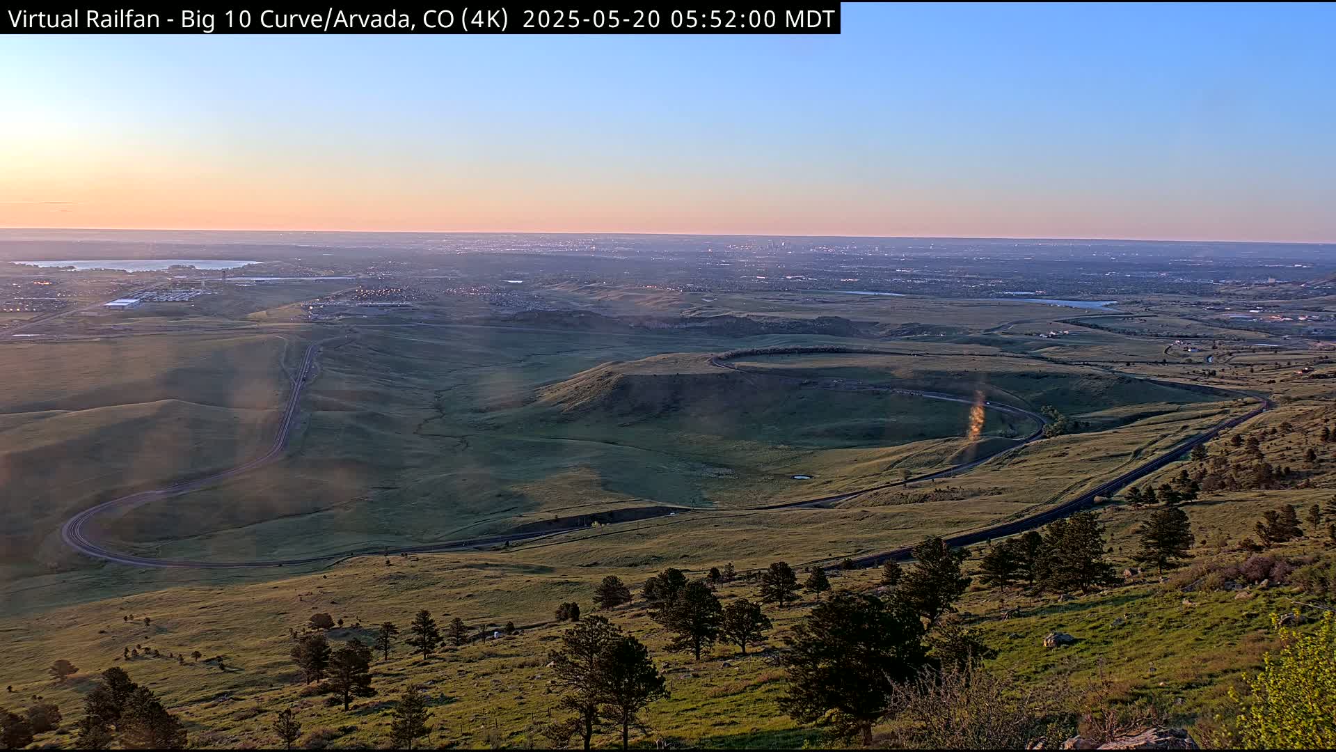 A sweeping vista of a curving road in a grassy landscape at sunrise, overlooking a distant city under a clear sky.