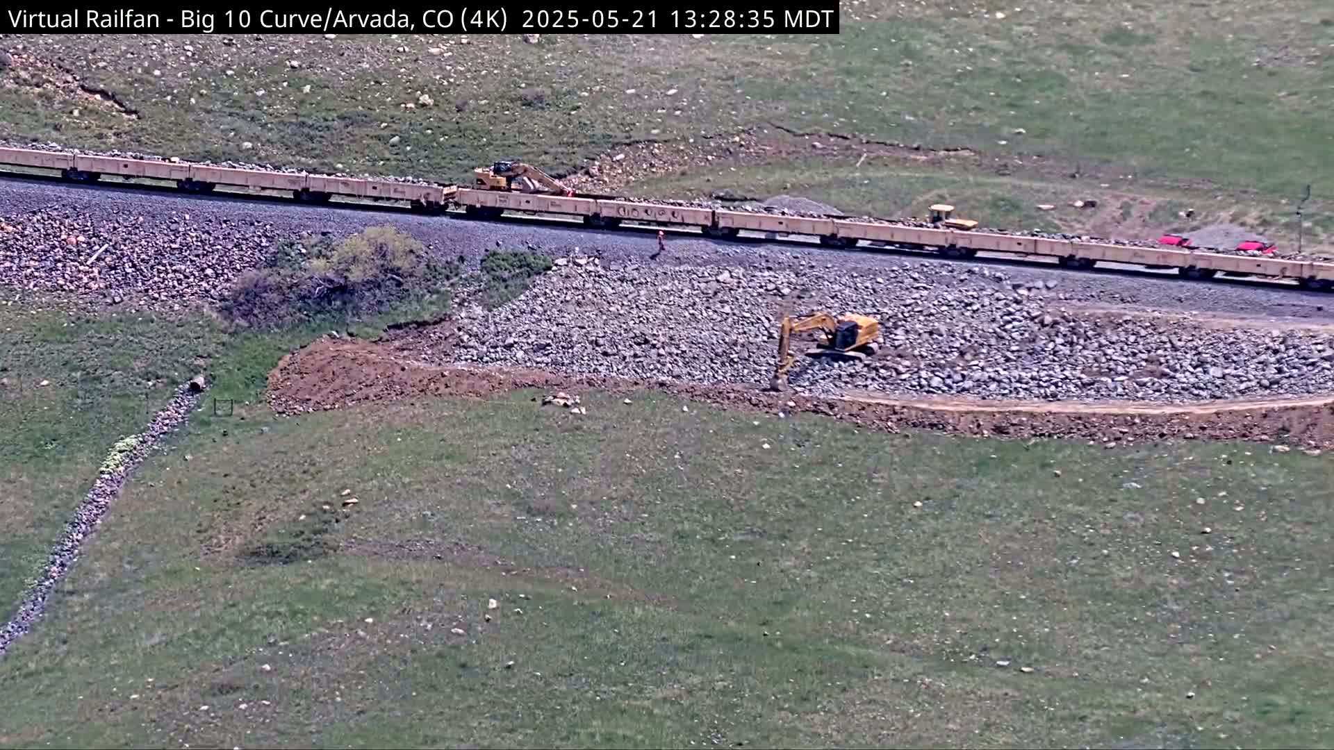 A long train carrying a load of rocks is parked near a construction site where excavators are working on a pile of rocks on a grassy hillside under a sunny sky.