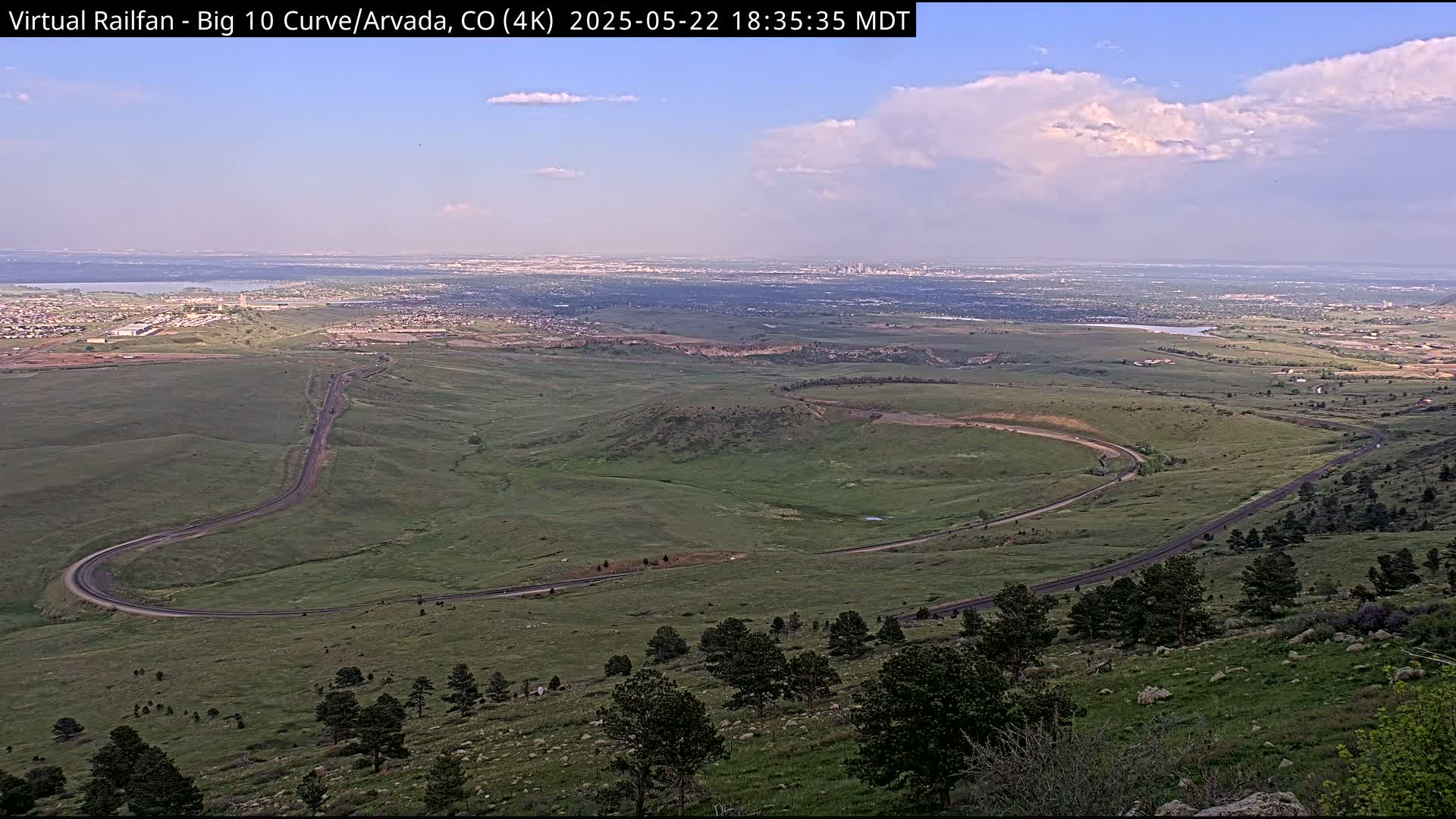 A winding road curves through a sprawling green landscape, overlooking a distant city under a partly cloudy sky.