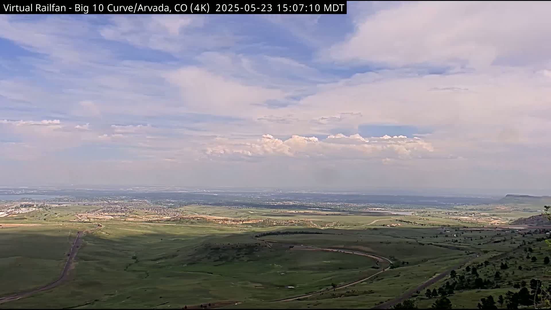 A high-angle view of a sprawling green landscape under a partly cloudy sky shows a distant city and a winding road leading down a hill.
