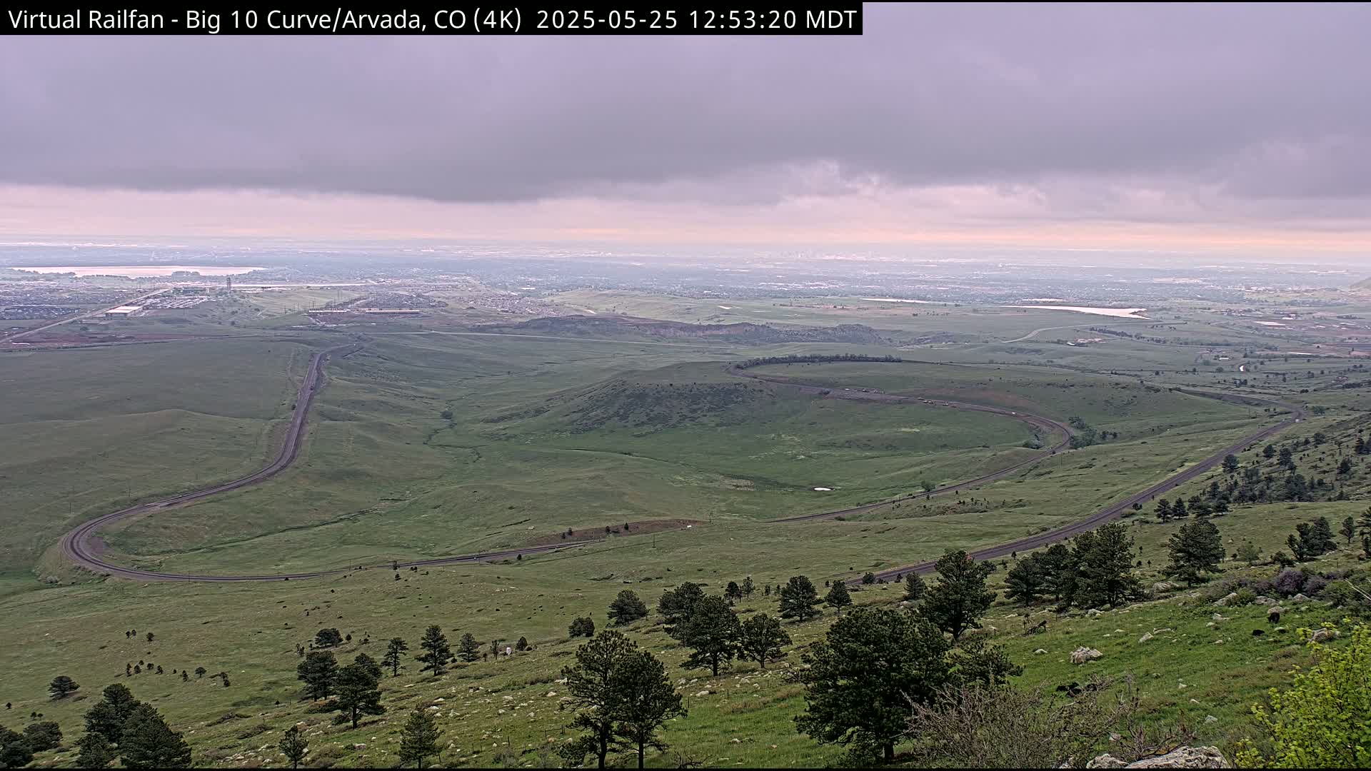 A winding road curves through a hilly, grassy landscape under an overcast sky, with a distant city visible on the horizon.