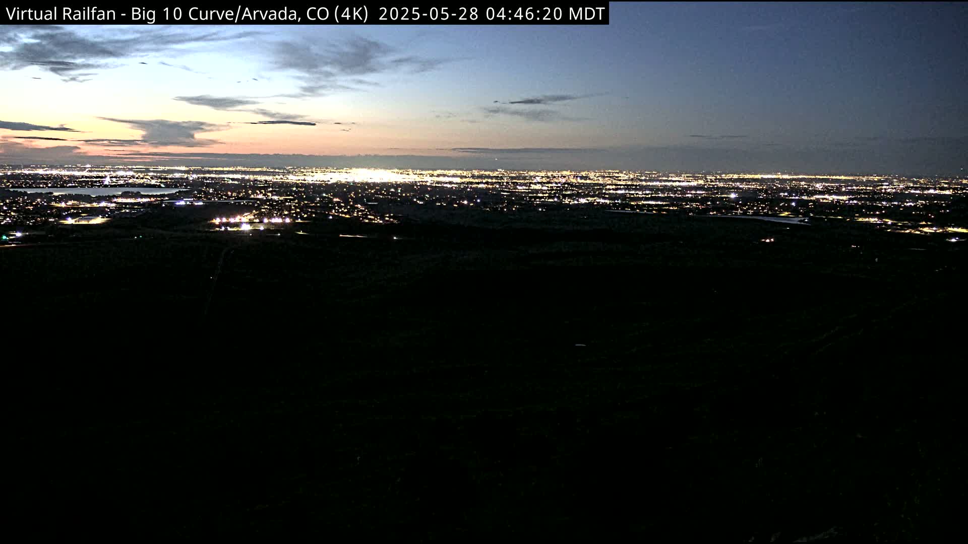 A nighttime, long-range view of a sprawling city illuminated by lights, with a dusky sky showing the remnants of a sunset.