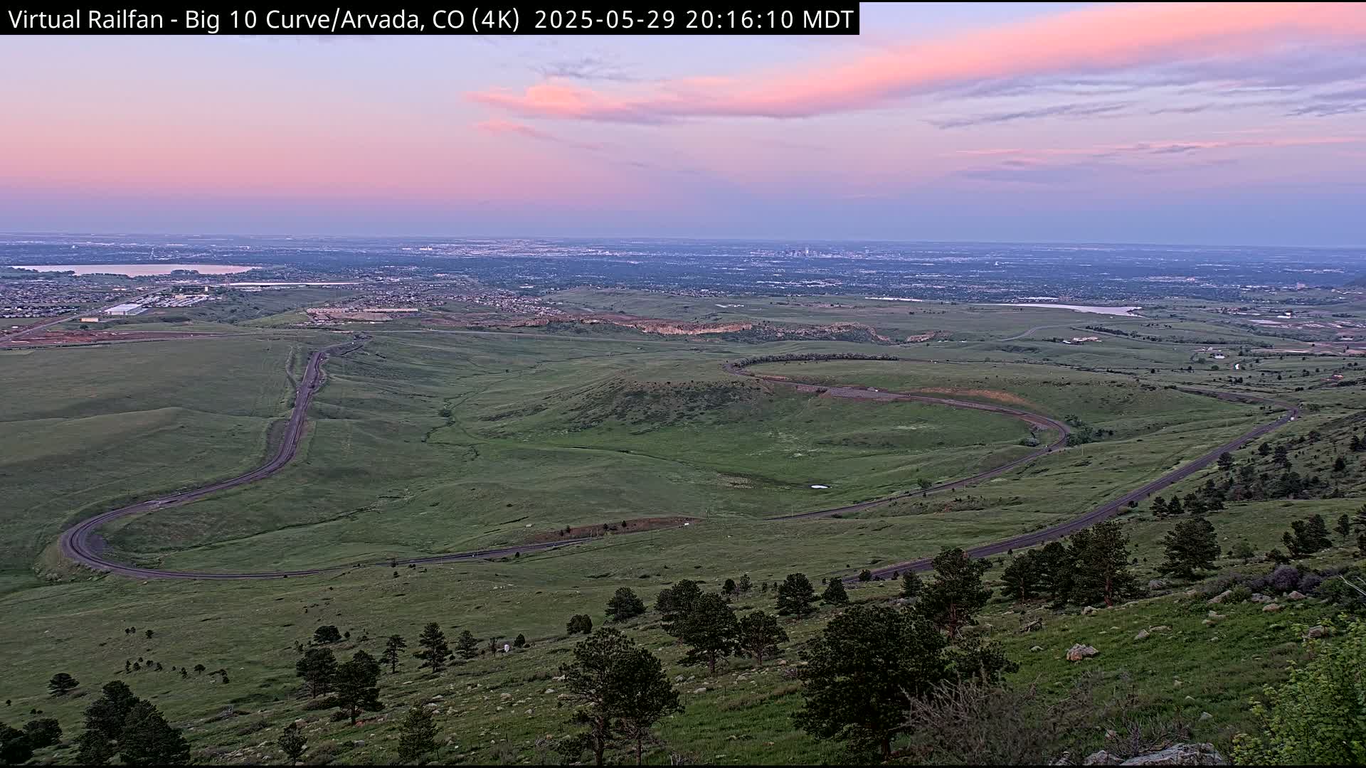 A winding road curves through a sprawling green landscape under a twilight sky, with a distant city skyline visible on the horizon.