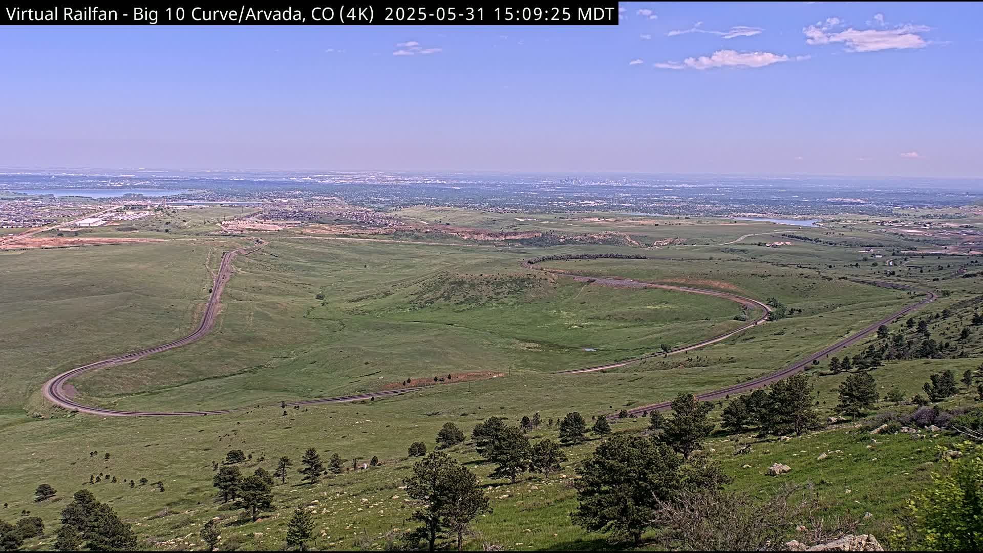 A high-angle, long shot on a partly sunny day shows a winding road curving through a sprawling green landscape, leading to a distant cityscape under a clear blue sky.