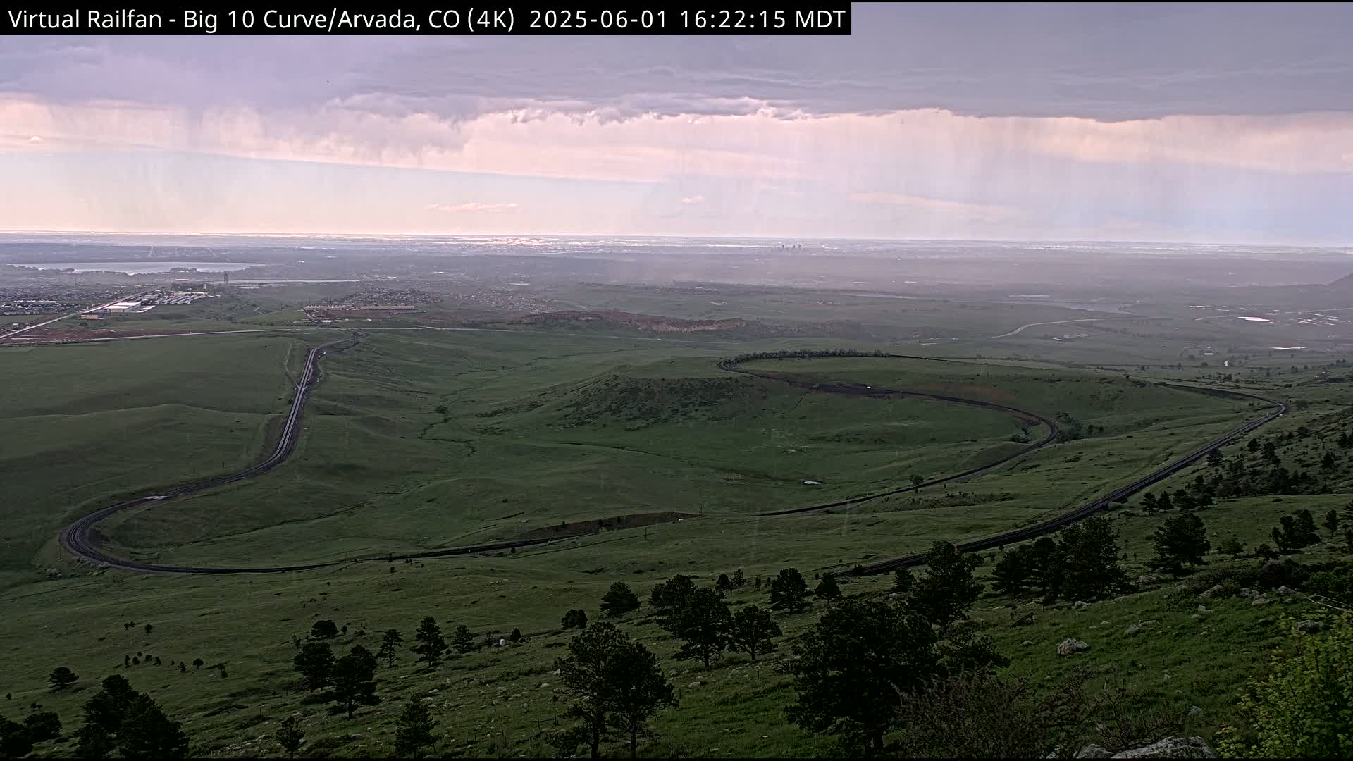 A winding road curves through a green, hilly landscape under a partly cloudy sky with light rain.