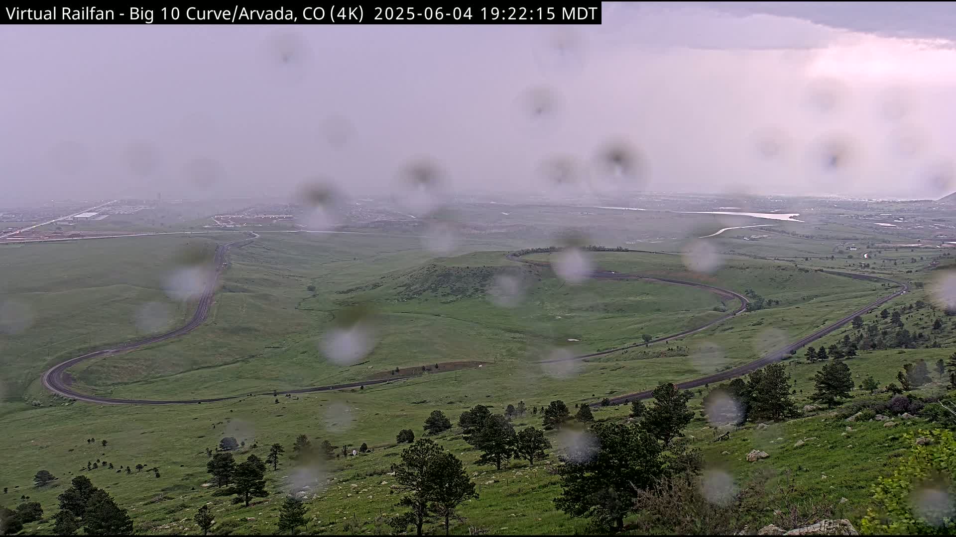 A winding road curves through a green, hilly landscape during a rainstorm.