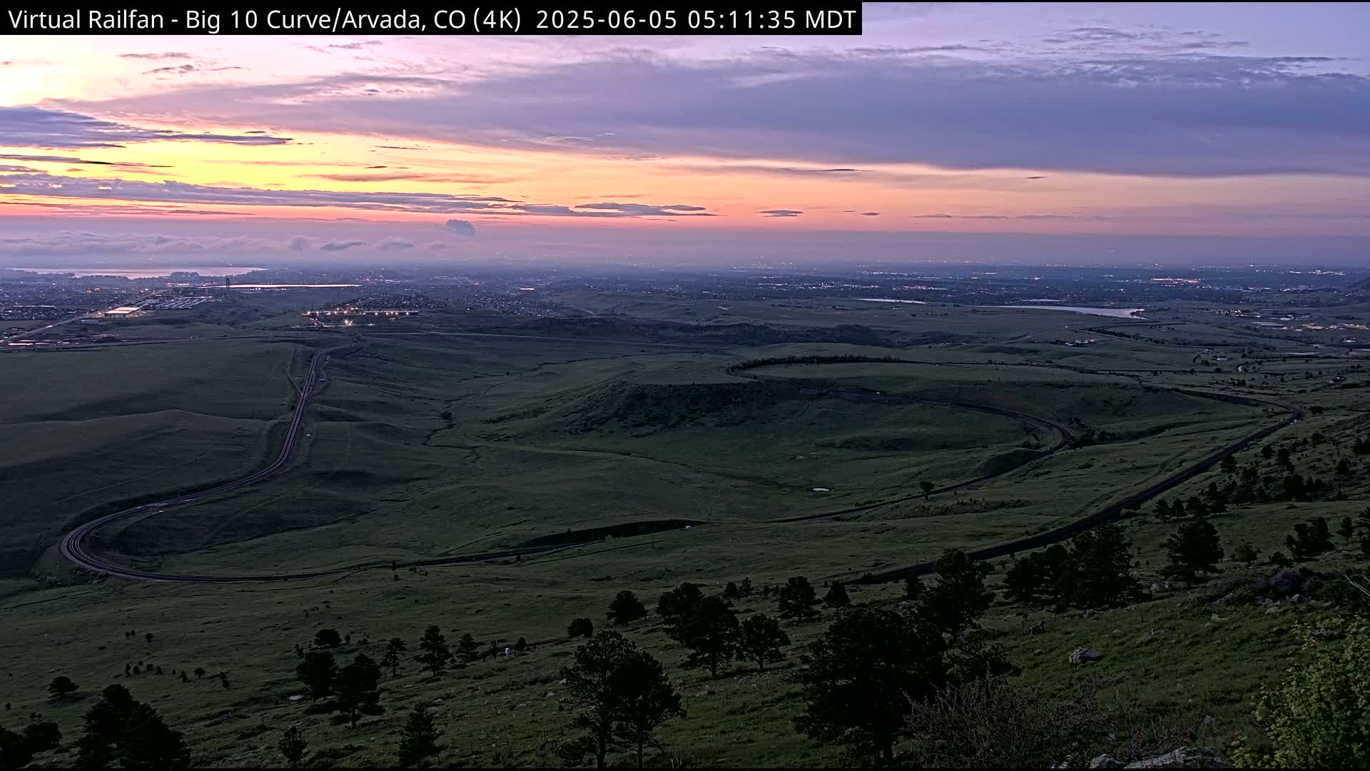 A winding road curves through a vast, green landscape under a twilight sky displaying soft pink and purple hues.