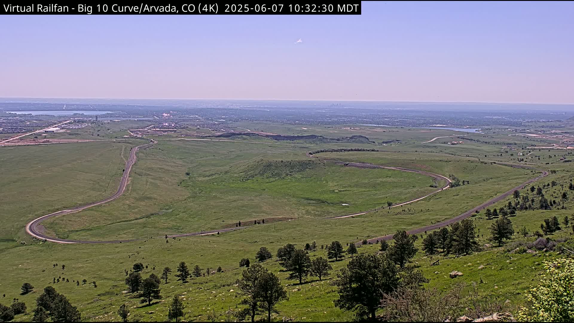 A winding road curves through a grassy landscape under a clear, bright sky, with a distant city skyline visible on the horizon.