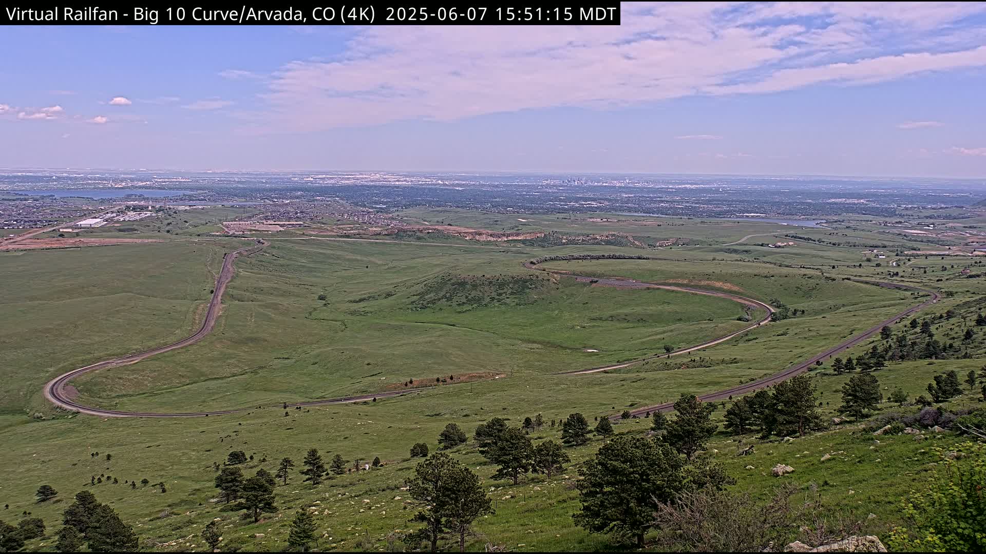A winding road curves through a grassy hillside, overlooking a distant city under a partly cloudy sky.