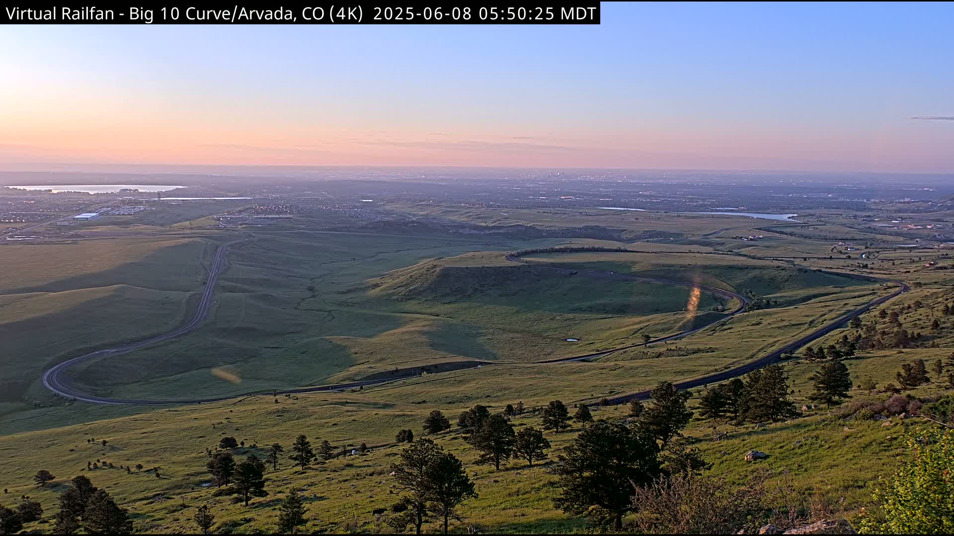 A winding road curves through a rolling, grassy landscape at sunrise, with a distant city skyline visible on the horizon under a clear, pale sky.