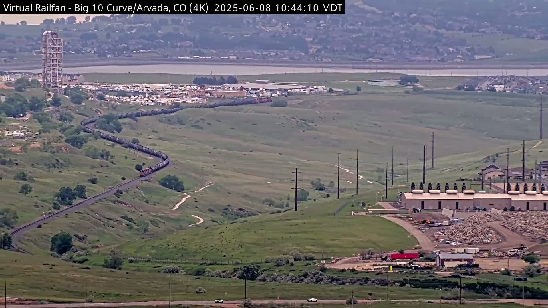 A long freight train curves through a grassy, hilly landscape under a partly cloudy sky, near industrial buildings and a body of water.