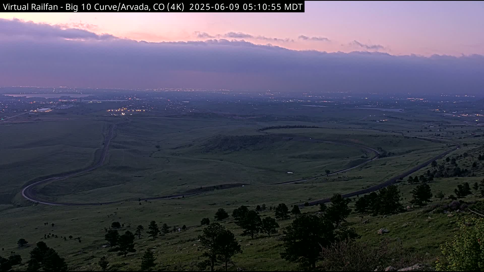 A winding road curves through a grassy landscape under a twilight sky, with a distant city faintly illuminated below.