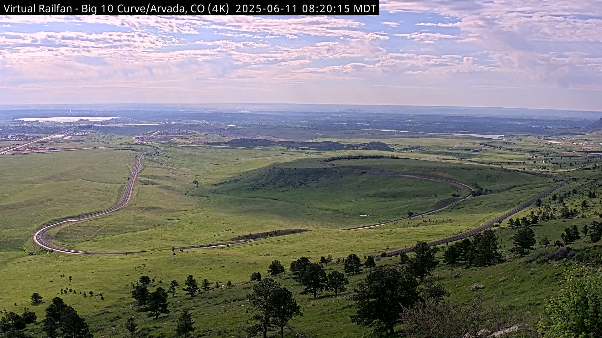 A winding road curves through a sprawling green landscape under a partly cloudy sky, with a distant city skyline visible on the horizon.
