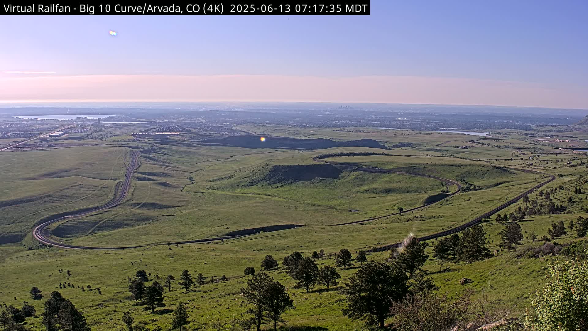 A winding road curves through a vast, green landscape under a clear, light-blue sky, with a distant cityscape visible on the horizon.
